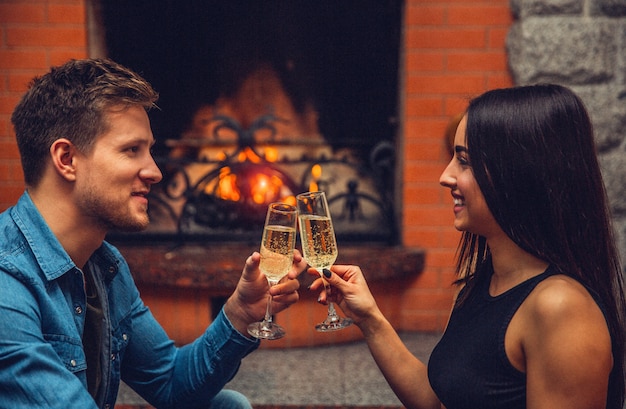 Cheerful young man and woman sit in front of each other and smile. They hold glasses with champagne and touch them. Couple smile. They sit at fireplace.