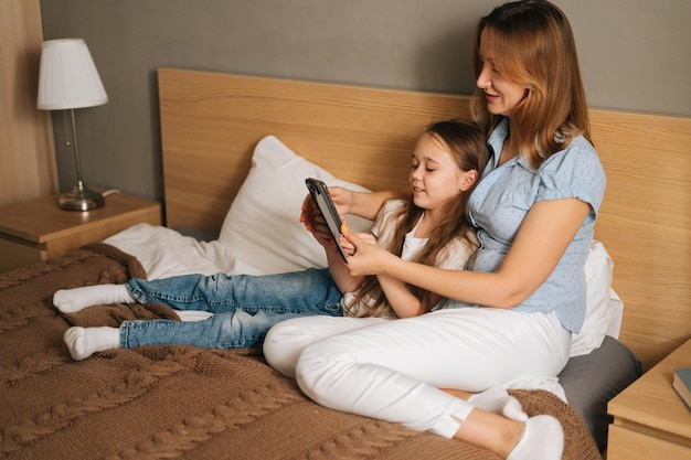 Cheerful young mother hugging to cute smiling daughter together watching fun video on mobile phone