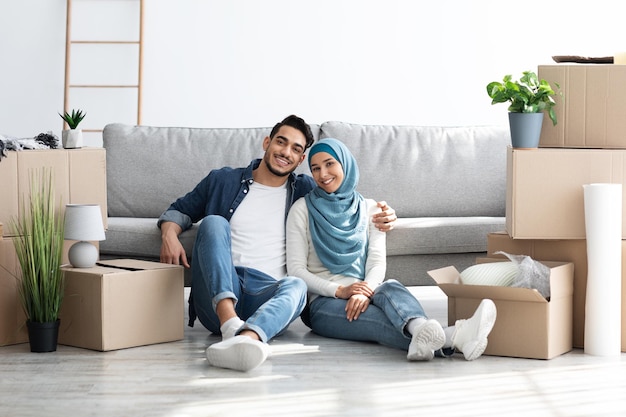 Cheerful young muslim family husband and wife in hijab posing while moving to their new apartment, sitting on floor by sofa among paper boxes with stuff, embracing and smiling at camera, copy space