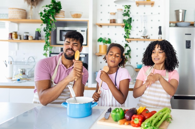 Cheerful young parents and little daughter having fun while baking together in kitchen happy African American family singing and fooling using spatula and whisk as microphones copy space