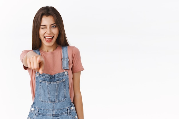 Cheerful young pretty girl in stylish outfing extand hand and pointing finger at camera, picking you, making confident choice with pleased smile, winking sassy, standing white background