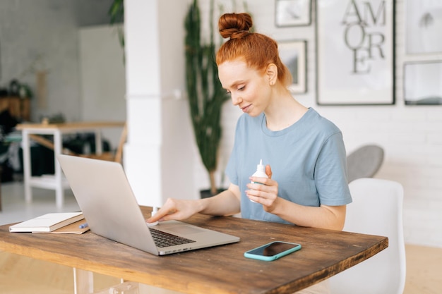 Photo cheerful young redhead woman is cleaning keyboard of laptop computer with sanitizer before starting work in light room at the home office. concept of remote work or education from home