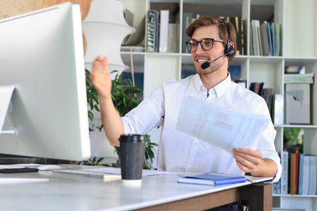 Cheerful young support phone male operator in headset at workplace while using computer