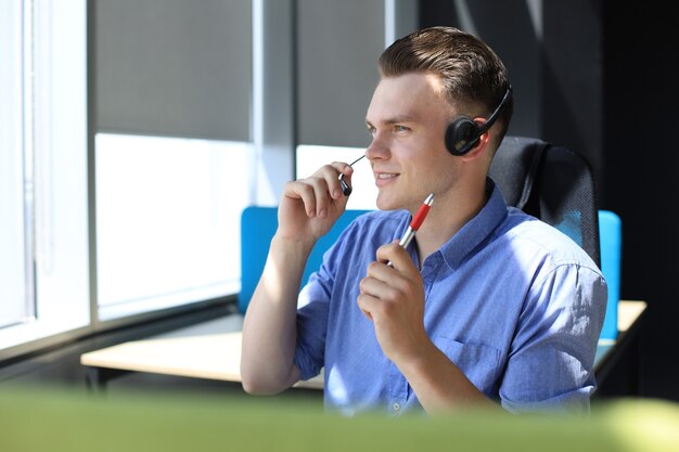 Cheerful young support phone male operator in headset, at workplace while using laptop, help service and client consulting call center concept.