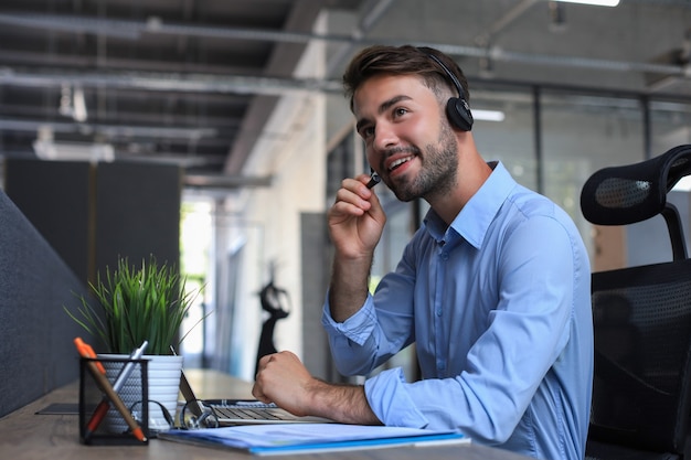 Cheerful young support phone male operator in headset, at workplace while using laptop, help service and client consulting call center concept.