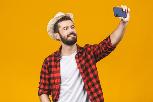 Cheerful young travel man in hat taking selfie with smartphone isolated over yellow wall.