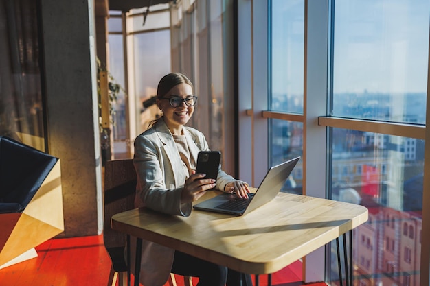 Cheerful young woman in casual wear having telephone conversation sitting at working place near laptop in office smiling female freelancer calling to colleague for share good news about business