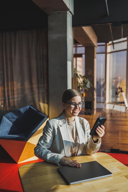 Cheerful young woman in casual wear having telephone conversation sitting at working place near laptop in office smiling female freelancer calling to colleague for share good news about business