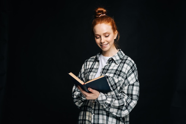 Cheerful young woman college student reading book on black isolated background