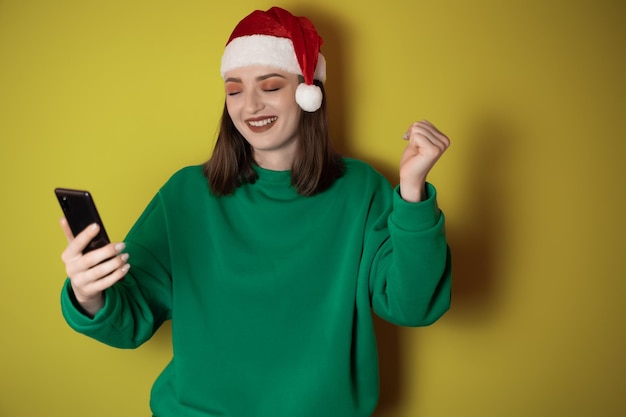 Cheerful young woman dressed in a green sweater and santa hat holding a mobile phone in her hand making a winner gesture isolated on a green background Happy new year 2023 holiday concept