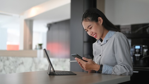 Cheerful young woman employee sitting in front of computer tablet at office desk and using smart phone.