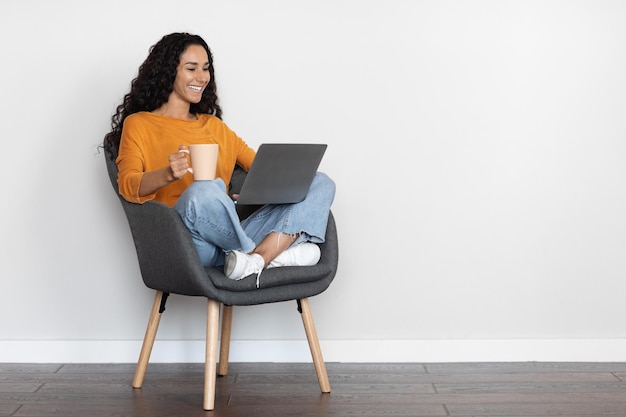 Cheerful young woman freelancer using laptop at home drinking coffee