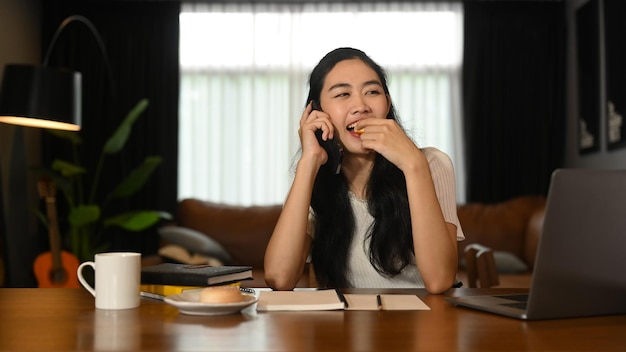 Cheerful young woman having pleasant phone conversation with her friend while sitting in cozy home office