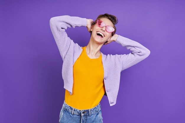 Cheerful young woman holding hand behind head while standing against purple background