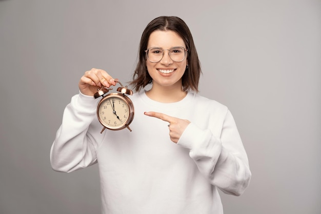 Cheerful young woman is pointing at the alarm clock she is holding