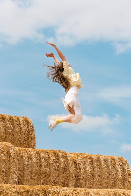 Cheerful young woman jumps from high haystack Happy girl has fun on the farm on blue sky background