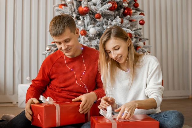 Cheerful young woman and man opening present boxes sitting on the floor near decorated christmas tree enjoying happy moments celebrating New Year's Eve