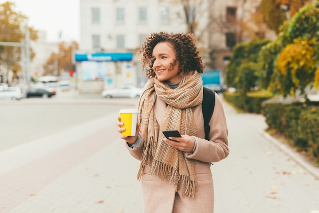 cheerful young woman outdoor during autumn walk on city streets and drinking cup of coffee