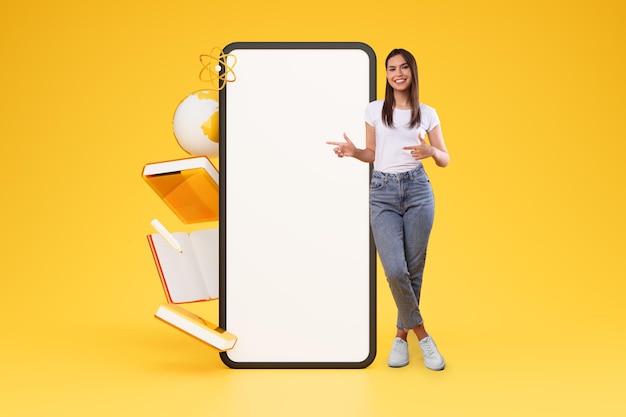 Cheerful young woman pointing at big phone over yellow background