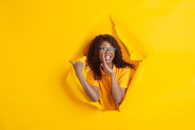 Cheerful young woman poses in torn yellow paper hole background, emotional and expressive