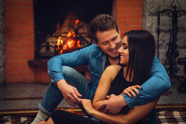 Cheerful young woman sit together with man on floor. He embrace her. They sit happily close to fireplace