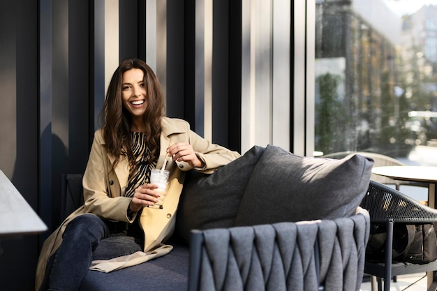 Cheerful young woman sitting on a sofa drinking a drink on a terrace in a cafe Attractive girl resting outdoors looking at camera