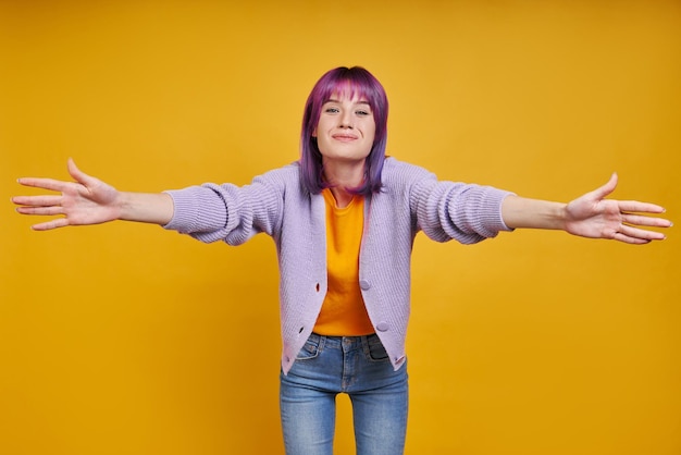 Cheerful young woman stretching out hands for hugging while standing against yellow background