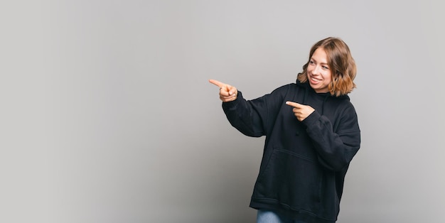 Cheerful young woman wearing black hoodie and pointing away at copyspace over grey background