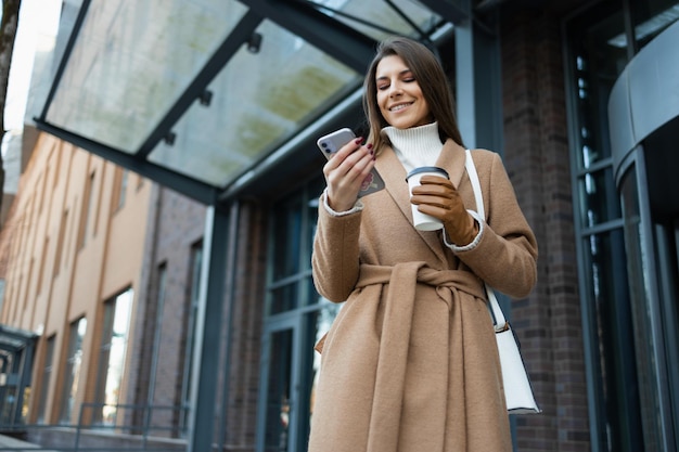 Cheerful young woman wearing brown coat standing in downtown holding paper coffee cup