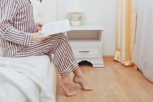 Cheerful young woman wearing pajamas is reading book in bed enjoying literature in nice bedroom Leisure activities and intelligent people concept