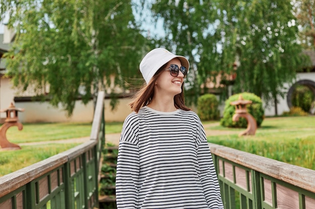 Cheerful young woman wearing striped shirt and panama relaxing on park standing on bridge enjoying summer weather smiling girl in sunglasses