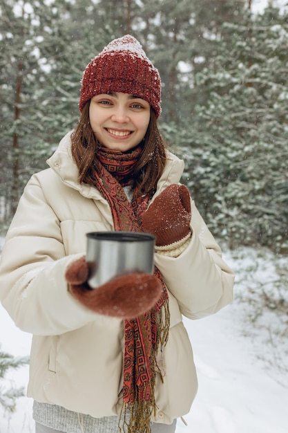 Cheerful young woman in winter outerwear with thermos cup of hot tea looking at camera