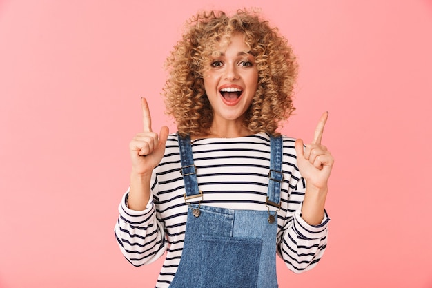 Cheerful young woman with curly hair wearing casual clothes, pointing up at copy space