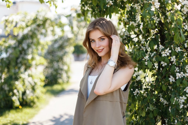 Cheerful young woman with a lovely smile with natural make-up with blue eyes in an elegant summer beige vest near the green flowering trees in the park. Joyful positive girl