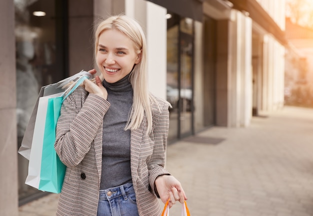 Cheerful young woman with paper bags smiling and looking away while standing on sidewalk on city street after shopping