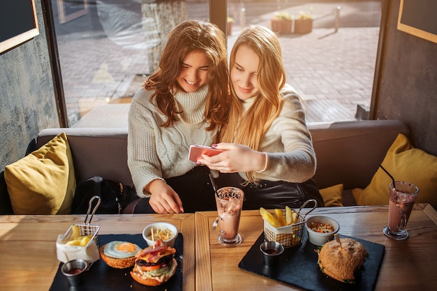 Cheerful young women look at phone together. Blonde model hold it in hands. They watch and smile. There are food at table. Young women sit in side at table.