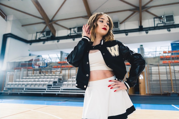 Photo a cheerleader wearing black and white uniform with a black jumper posing in a sports hall the stands