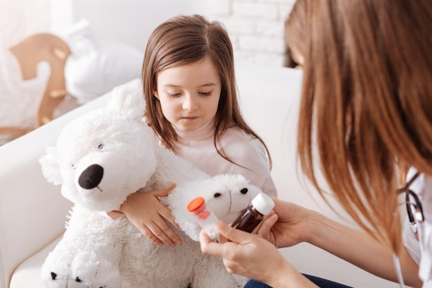 Cheerless little girl holding her fluffy toy and going to take pills while professional doctor giving her tipps