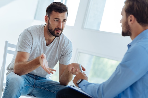 Cheerless moody young man looking at his therapist and asking for advice while looking for a solution to his problems