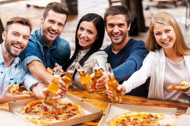 Cheers to you! Group of happy young people bonding to each other and stretching out bottles with beer while standing outdoors