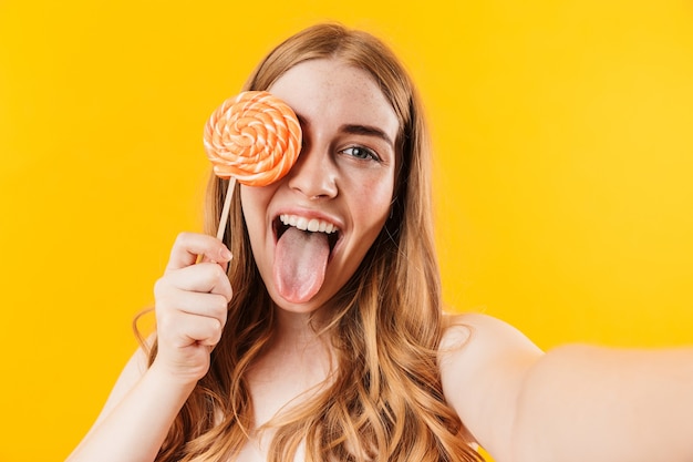 a cheery pleased happy young cute teenage girl posing isolated over yellow wall holding sweet candy lollipop take selfie by camera showing tongue.