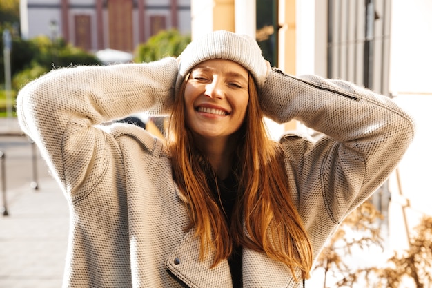 Cheery young woman dressed in autumn coat and hat walking outdoors at the city street