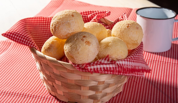 Cheese bread, basket lined with red and white fabric filled with cheese bread on a checkered towel and a white cup.