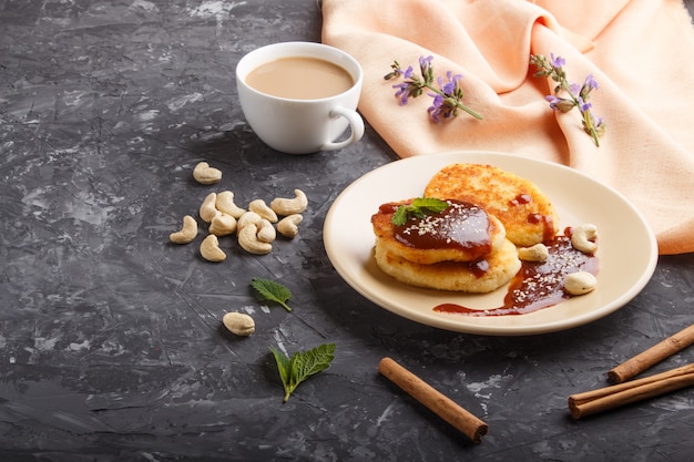 Cheese pancakes with caramel sauce on a beige ceramic plate and a cup of coffee on black concrete