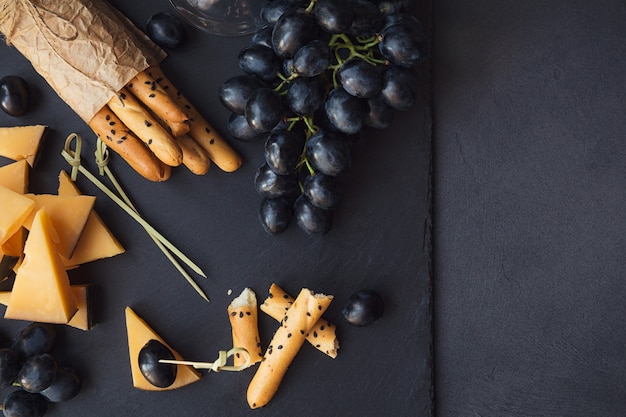 Cheese plate served with crackers, grapes and glass of white wine on dark background. Old gouda cheese on tasting plate. Top view with copy space.