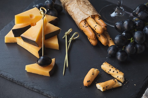 Photo cheese plate served with crackers, grapes and glass of white wine on dark background. old gouda cheese on tasting plate