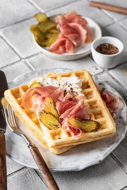 Cheese waffles with ham cream cheese and cucumber with cutlery and linen napkin on gray tile background Delisious savory breakfast