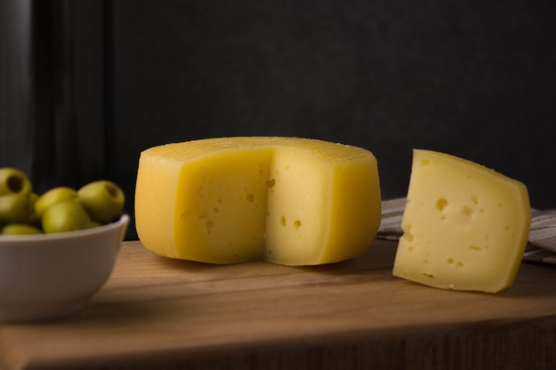 Cheese wheel with a cut piece on a wooden Board. Green olives nearby