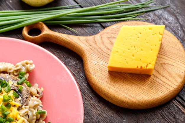 Cheese on a wooden board next to green onions on the table and a fragment of a plate with pasta in a creamy sauce