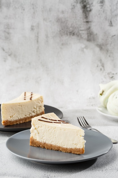 Cheesecake New York or classic cheesecake with cup of coffee on white table. Closeup view. Tasty breakfast. Piece of cake on black plate, white cup on white marble background. Vertical photo.